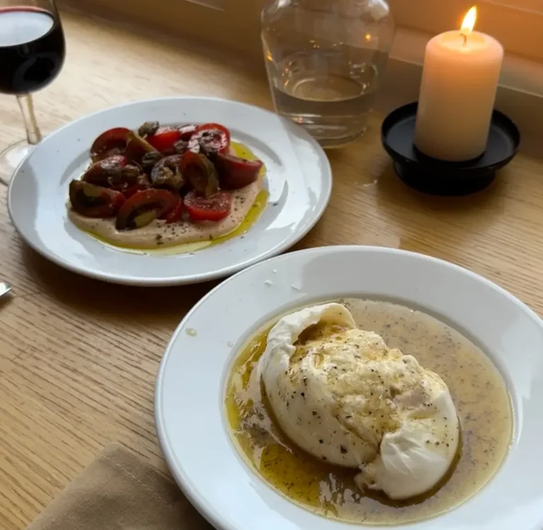 a table set with plates of food and a candle. The setting is in Darling's Eatery and Studio, a restaurant in East London known for Italian cuisine. The specific dish shown is burrata served with truffle olive oil and heritage Sicilian tomato salad.