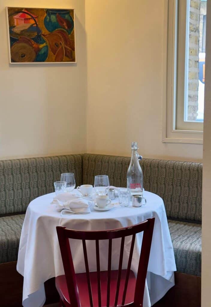 Elegant bistro table with a white tablecloth, empty glassware, cups, and a bottle of sparkling water. A red chair and abstract wall art complete the setting in this charming restaurant in London.