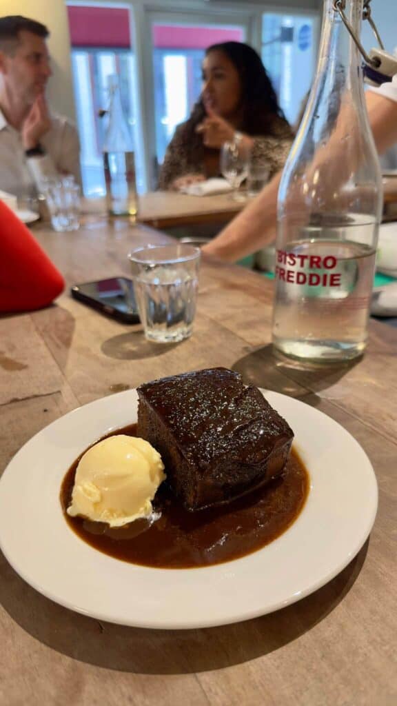 A square slice of sticky toffee pudding with a rich, glossy caramel sauce, served alongside a scoop of vanilla ice cream on a white plate. The background shows a clear bottle labeled “Bistro Freddie” and a few other diners blurred out, giving the image a lively, casual dining ambiance