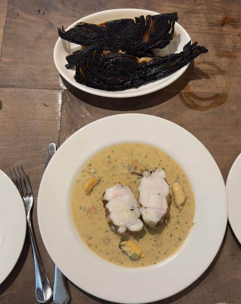 A plate featuring two pieces of monkfish served in a creamy, herb-infused sauce with a few mussels scattered around. In the background, there’s a small bowl with charred cabbage, heavily blackened on the edges, adding a contrast to the dish.