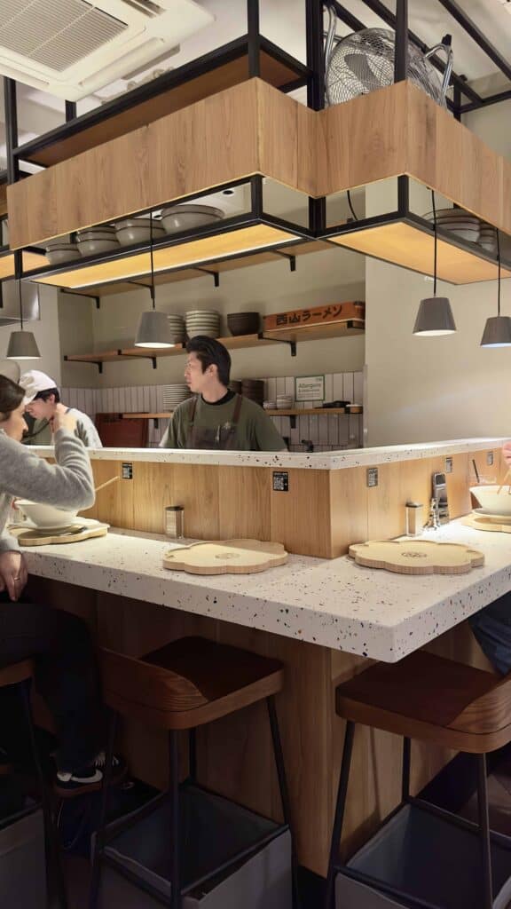 Interior of Ramen Moto London featuring a Japanese-style counter dining setup, wooden stools, and an open kitchen where chefs prepare ramen.