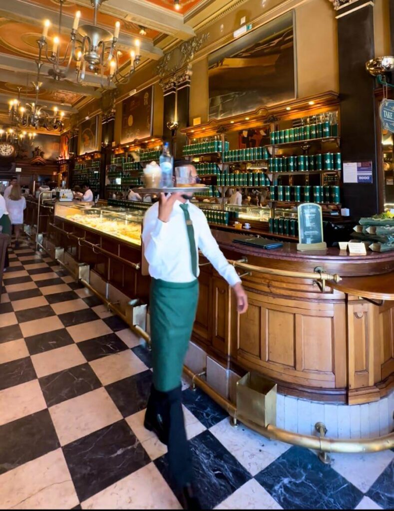 A young waiter in a white shirt and green apron walks through A Brasileira, one of Lisbon's oldest coffee shops, with a tray serving drinks