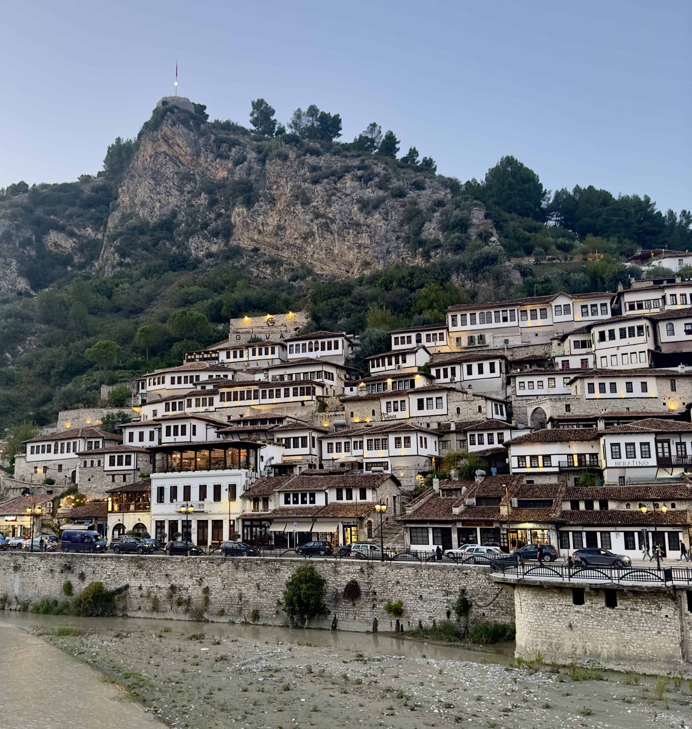 Sunset view of Berat, Albania, with traditional Ottoman-style buildings cascading down the hillside. Warm evening light illuminates the white stone walls and terracotta rooftops, creating a picturesque scene.