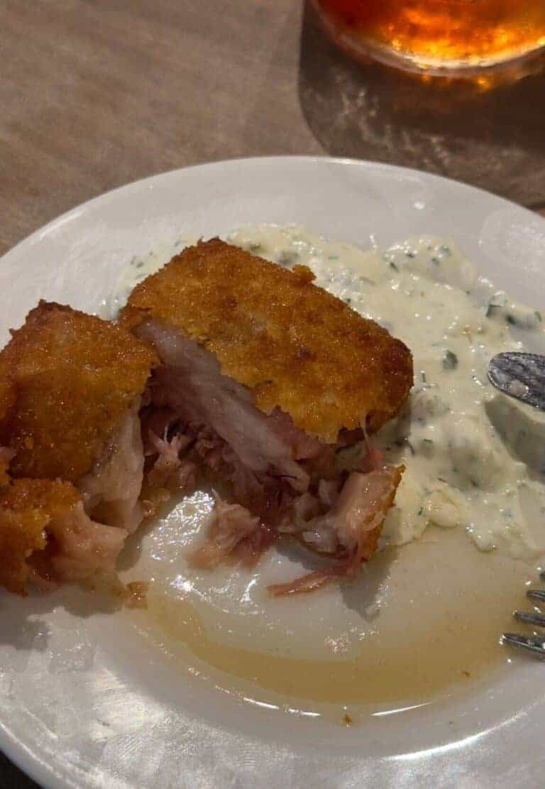 A close-up of a crispy pig head croquette split open to reveal tender, juicy meat inside. The croquette is served with a dollop of creamy tartare sauce on a white plate, with a glass of a reddish cocktail blurred in the background.