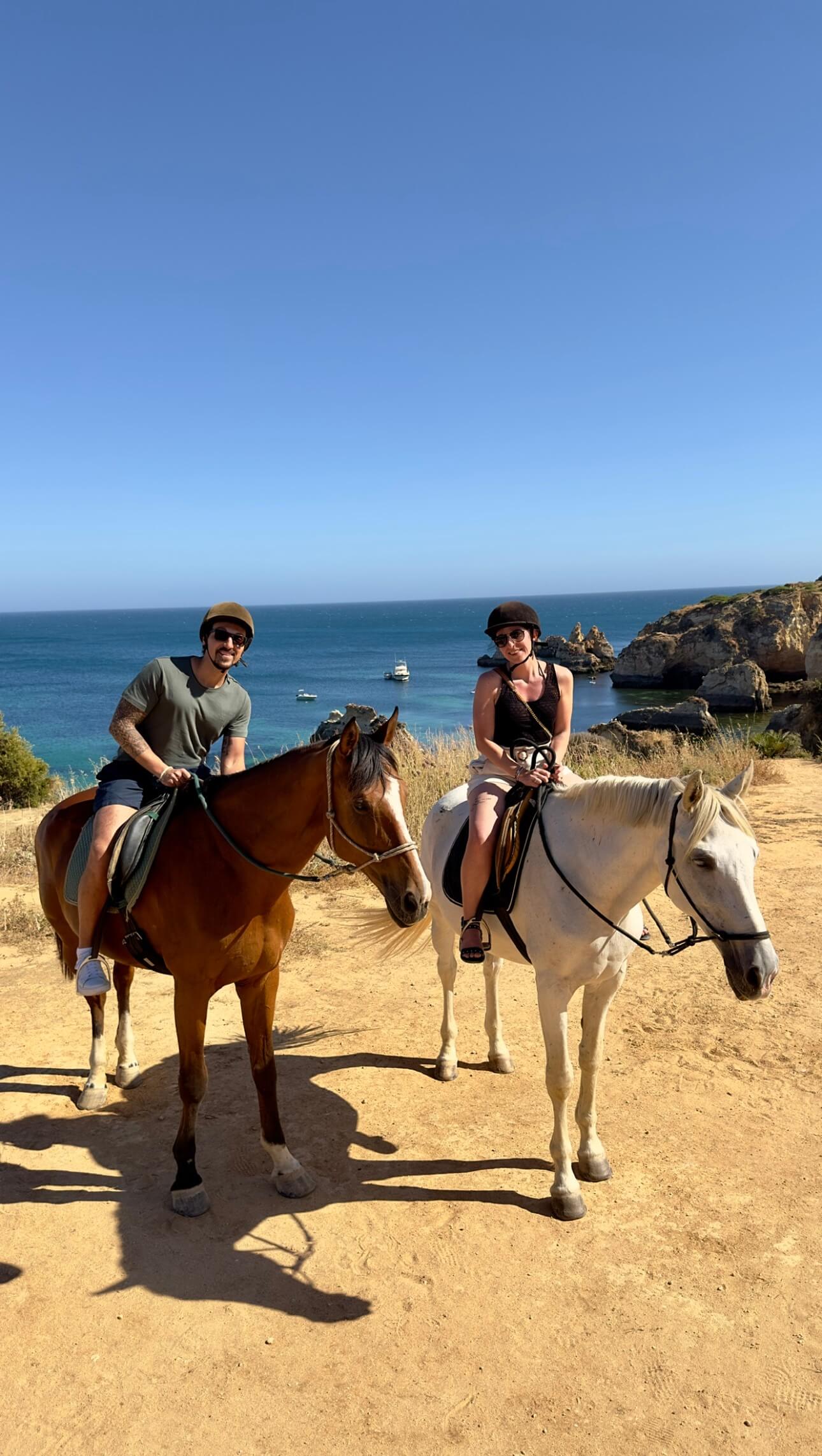 My husband and I riding a horse in the costline of Algarve, Portugal, with the background of the sea, boats and clear sky.