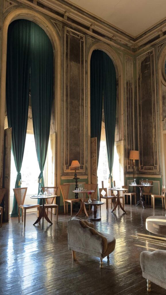 The grand interior design of Palacio do Grilo showing high ceilings, distressed walls, large arched windows with green curtains, and elegant dining tables
