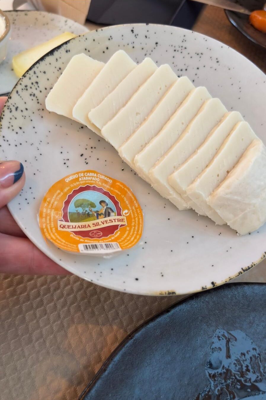 A hand holding a speckled plate of sliced Queijaria Silvestre cured goat cheese served as a traditional starter at Avô Casimiro restaurant in Portimão.