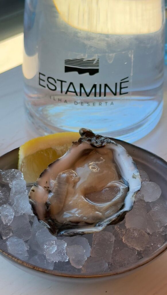 A fresh raw oyster served on a bed of crushed ice with a lemon wedge in front of an Estaminé Ilha Deserta water carafe, highlighted in an Algarve food guide.