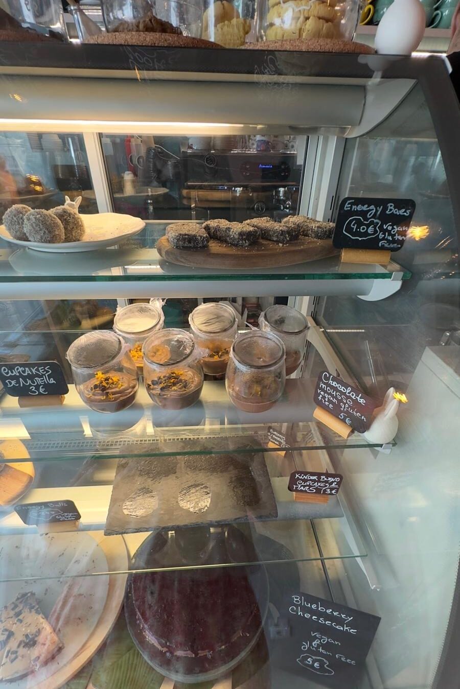 A glass bakery display case inside Sugar Rabbit Kaffé filled with vegan and gluten-free treats, including chocolate mousse jars, energy bars, cupcakes, and a blueberry cheesecake with handwritten price signs.
