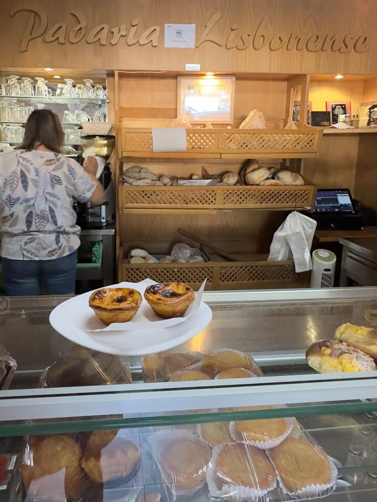The wooden service counter and pastry display case inside Padaria Lisbonese, showcasing two fresh pastel de nata tarts on a plate and rows of traditional bread loaves behind the worker.