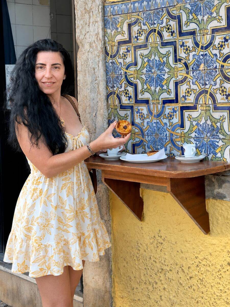 Blog author Sara holding a pastel de nata tart at a small wooden outdoor wall counter next to beautiful blue and white Portuguese tiles at Padaria Lisbonese bakery.