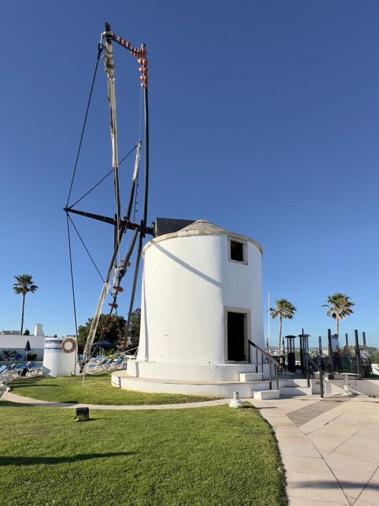 The exterior of the historic 1938 white windmill housing the Windmill restaurant in Albufeira, featuring its large traditional sails against a bright blue sky.