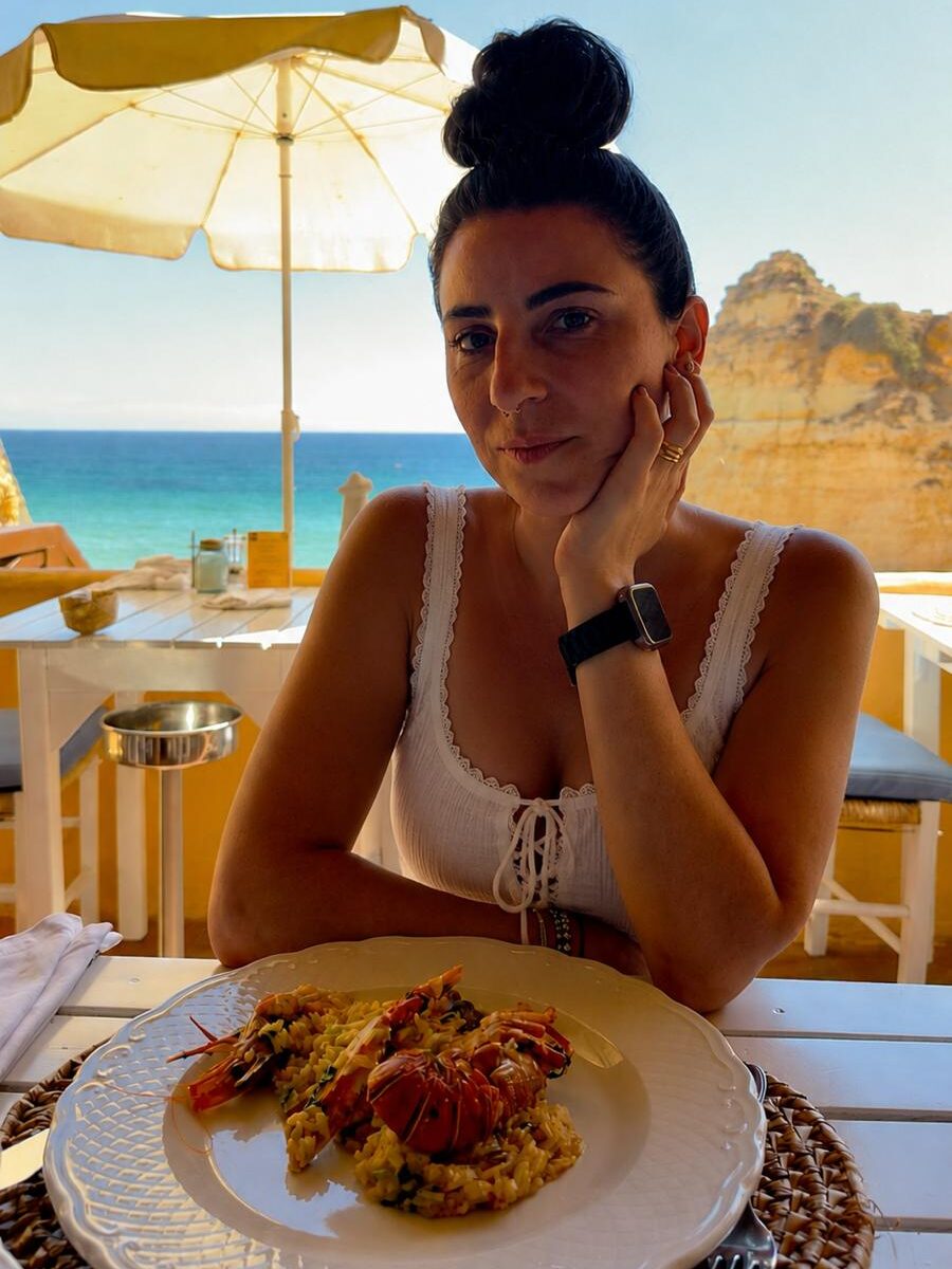 Blog author Sara sitting at a white wooden dining table under an umbrella at Caniço restaurant, smiling behind a plate of traditional Portuguese lobster and prawn seafood rice, highlighted in an Algarve food guide.