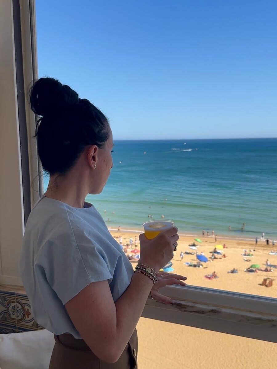 Myself standing by an open window at Sal Rosa bar in Albufeira, holding a premium cocktail and looking out over a crowded beach and the infinite blue Atlantic ocean.