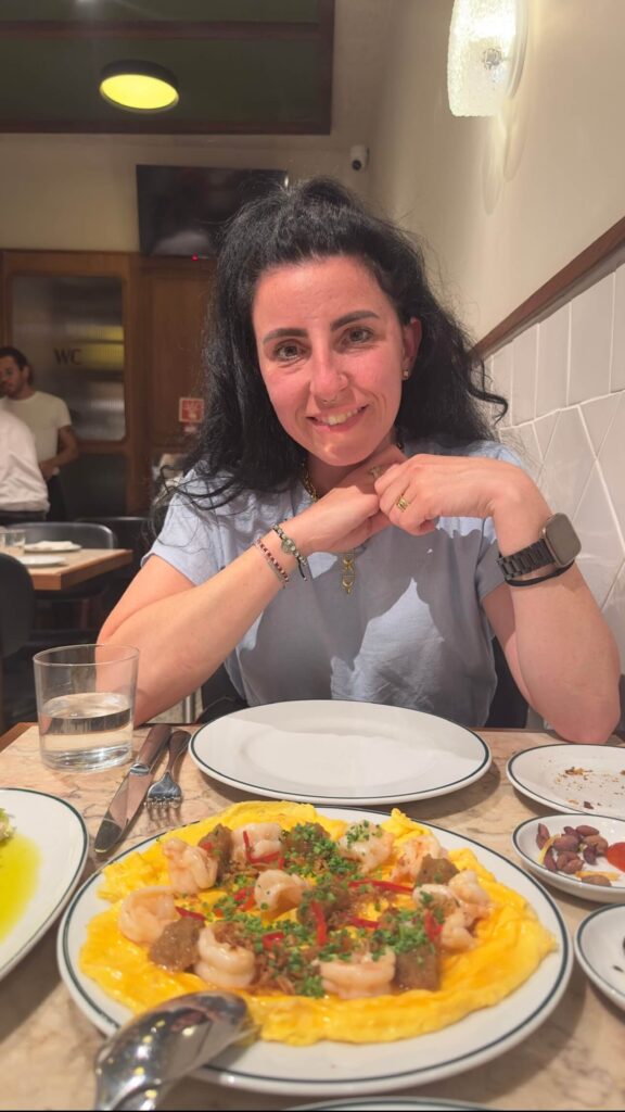 Blog author Sara sitting at a dining table inside Canalha, smiling in front of an open face omelet topped with prawns, fresh chives, and crisp onions, featured in a guide to the best restaurants in Lisbon.