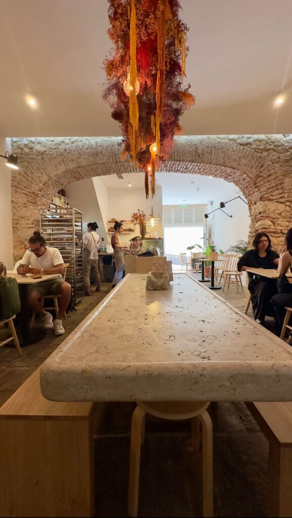 he rustic-chic interior of Fora bakery featuring a large stone communal table, stone archways, and a vibrant hanging dried flower installation above the seating area, included in a Lisbon food guide