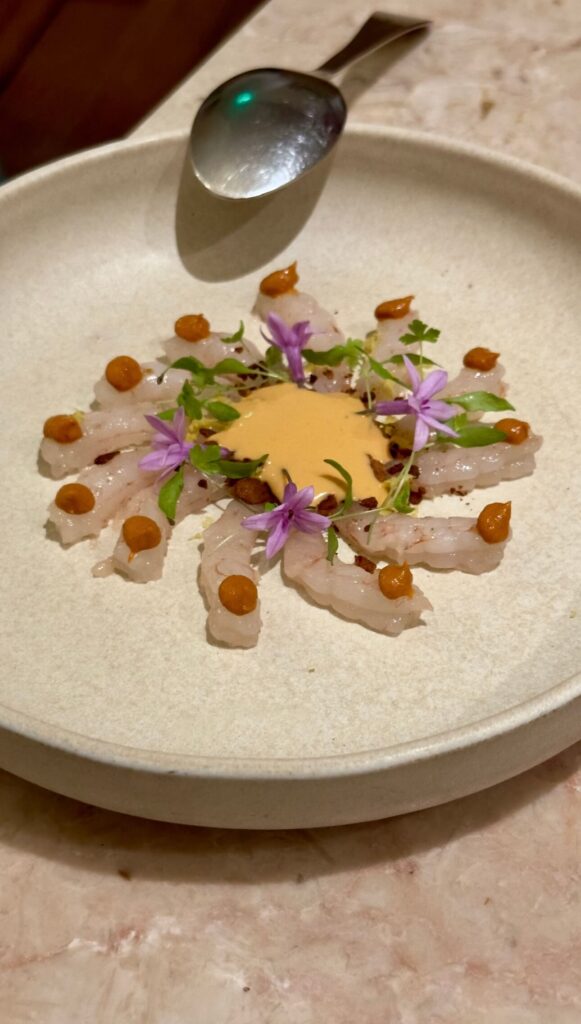 An elegantly plated raw shrimp prawn dish arranged in a circle, garnished with edible purple flowers, microgreens, and a central sauce at Ofício restaurant, featured in a Lisbon food guide