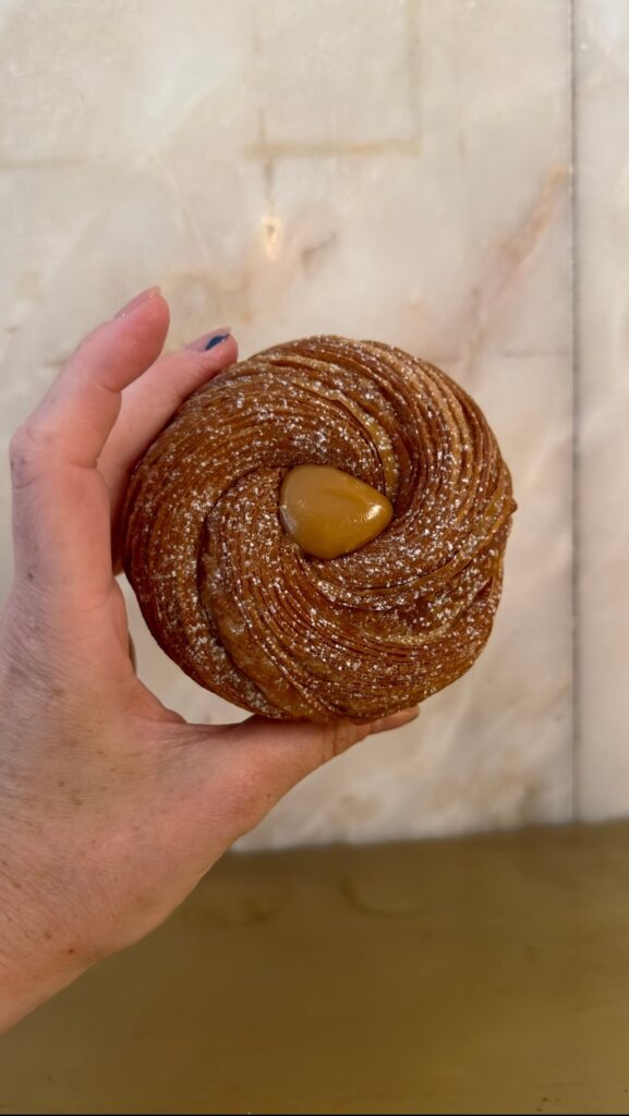 My hand holding a flaky, sugar-dusted caramel cruffin filled with a dollop of rich caramel in the center from Lully Parisian bakery, highlighted in a Lisbon food guide