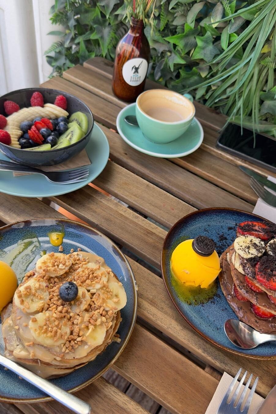 A wooden outdoor table spread at Sugar Rabbit Kaffé in Albufeira, displaying a fresh fruit bowl, a cup of coffee, Pea-Bananuts pancakes, and vegan No-tella pancakes with a side of sorbet against a green leaf wall.