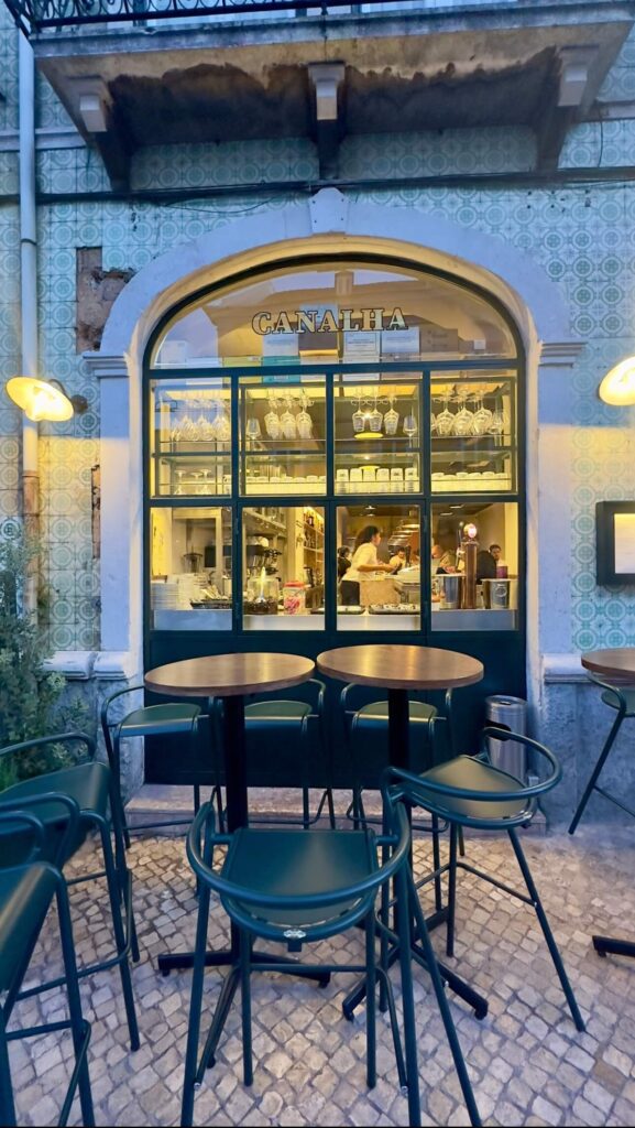 The charming tiled exterior of Canalha restaurant at twilight, featuring high bar tables, green stools, and a warm glow through the large arched window.