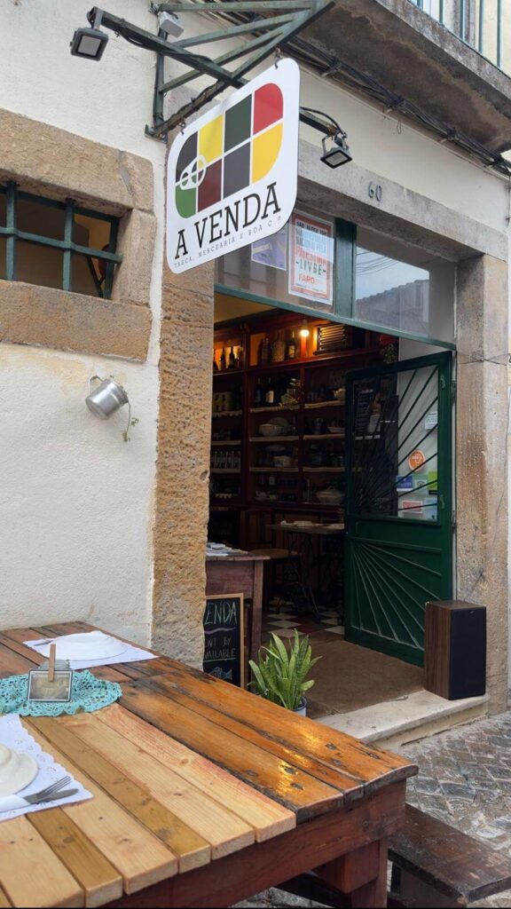 The charming street entrance and outdoor wooden tables of A Venda restaurant in Faro, featuring its distinctive colorful signboard hanging above an open green door.