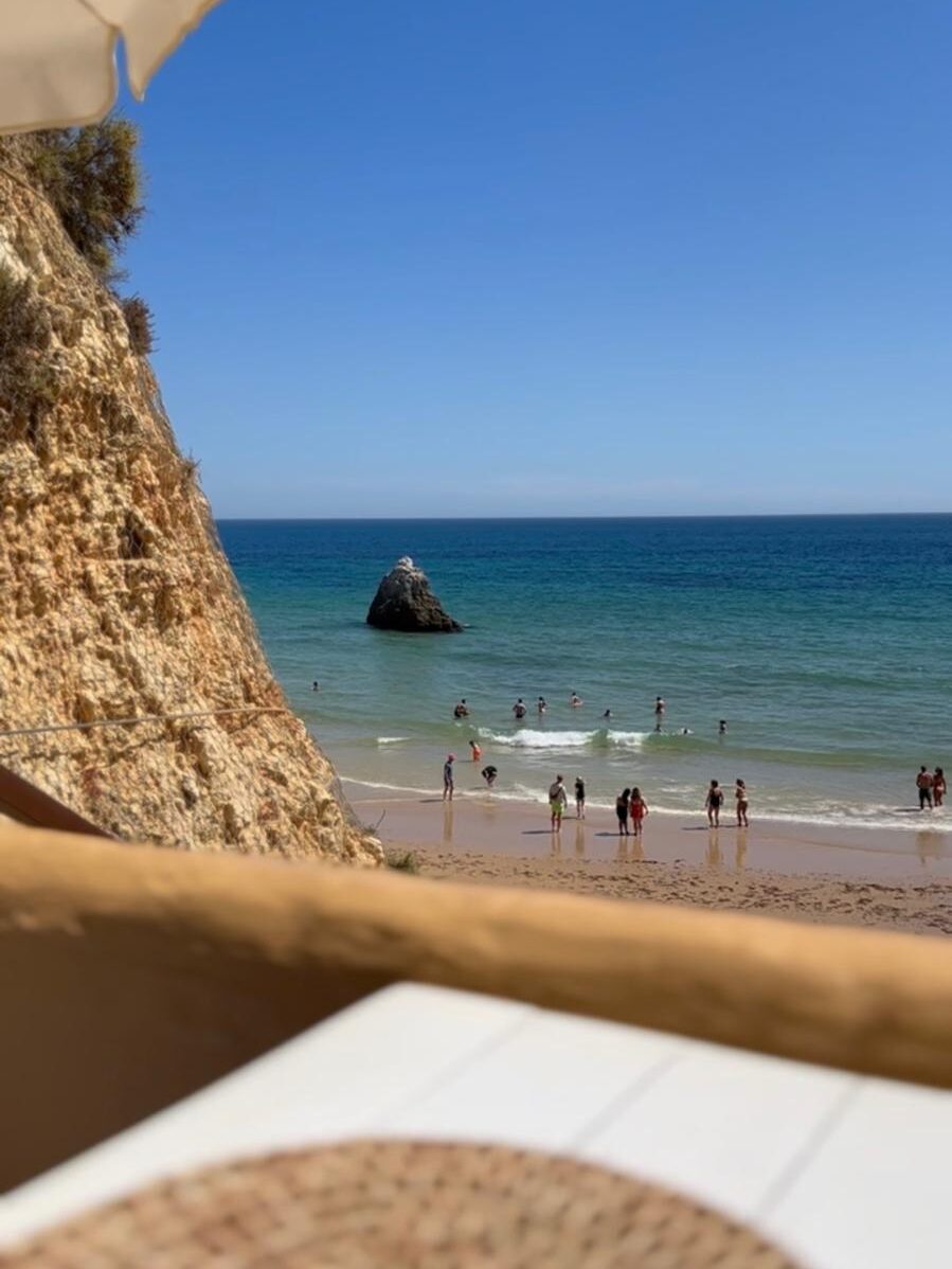 The stunning ocean view from an outdoor dining table at Caniço restaurant in Alvor, looking out over a sandy beach, clear blue water, and rugged cliffs, featured in a guide on where to eat in the Algarve.