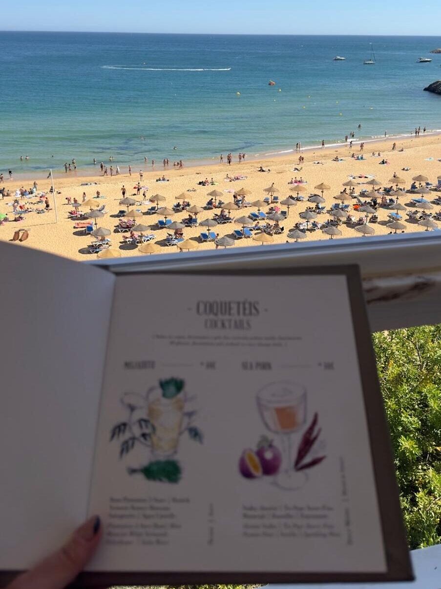 My hand while holding open the artistic sketchbook menu of Sal Rosa cocktail bar, showing illustrated drink recipes against a stunning background of Albufeira beach.