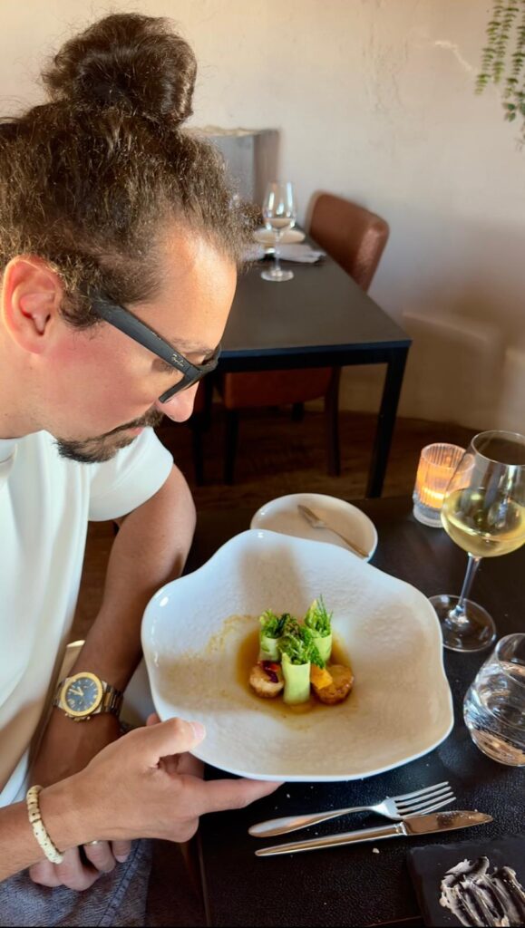 My husband admiring an artfully plated gourmet dish with fresh greens and seafood, served with a glass of white wine as part of a three-course tasting menu.